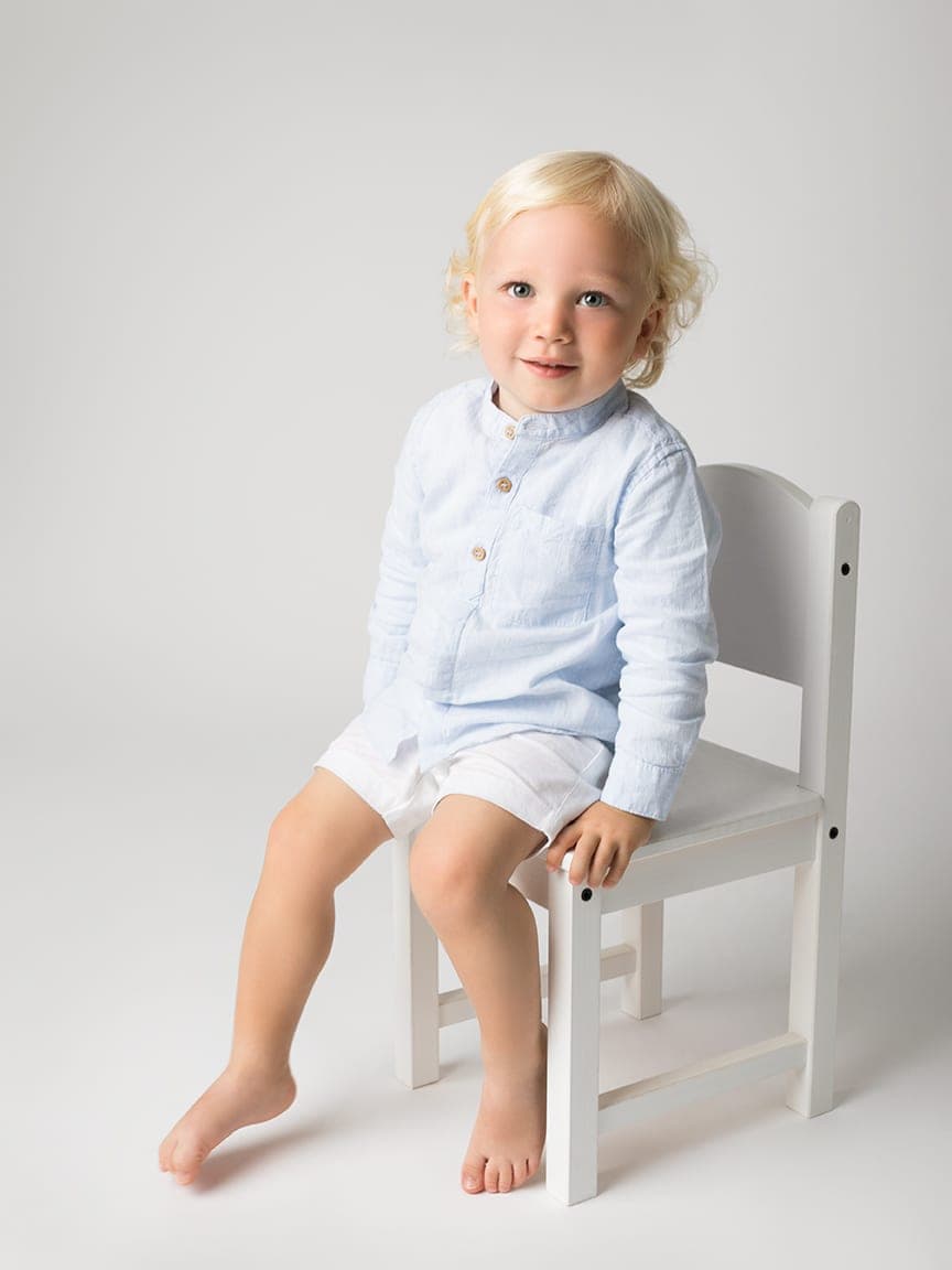 Little boy in a pale blue shirt sitting on a small white chair in the studio