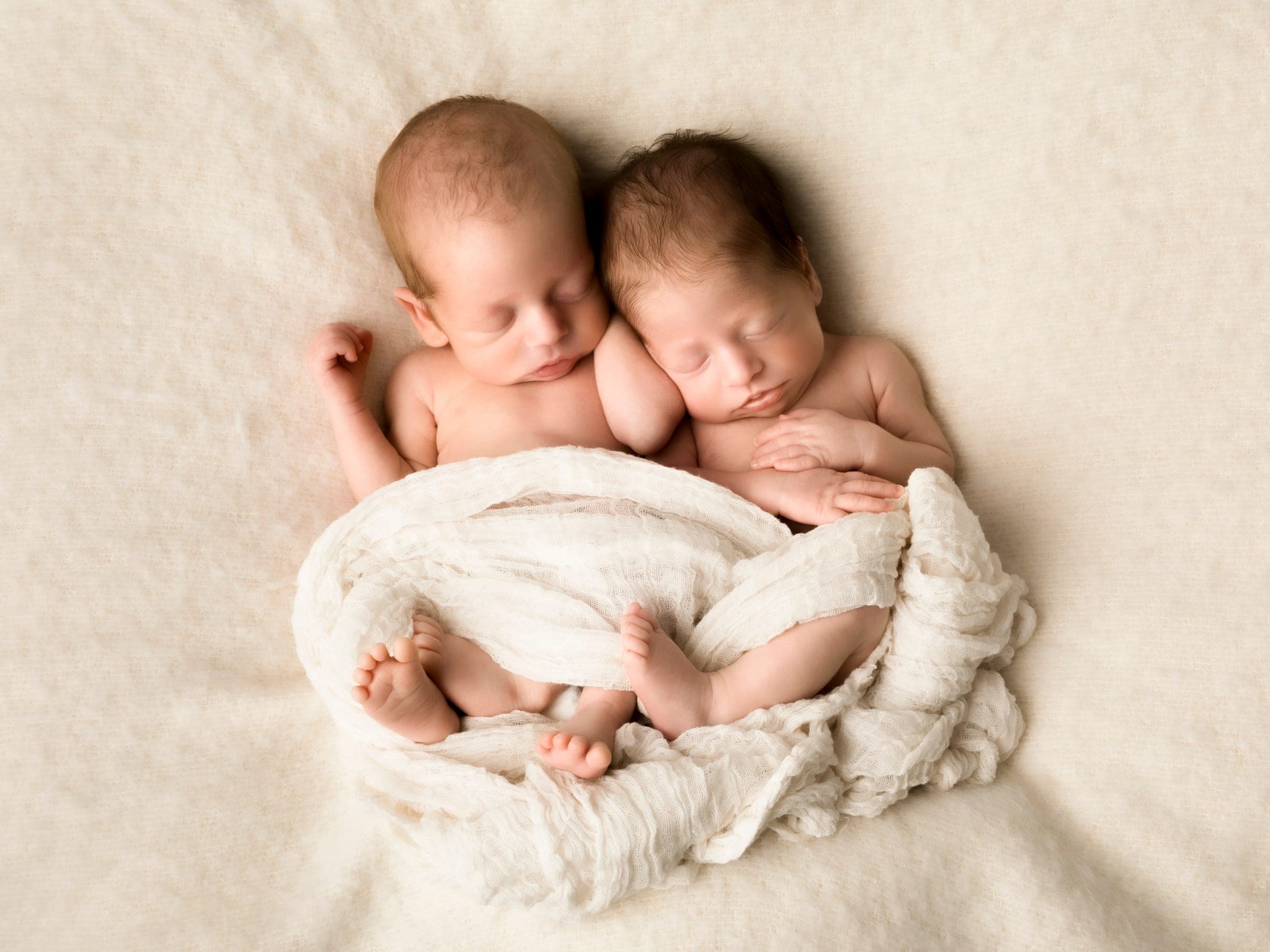 Newborn twins asleep side by side under a cream wrap