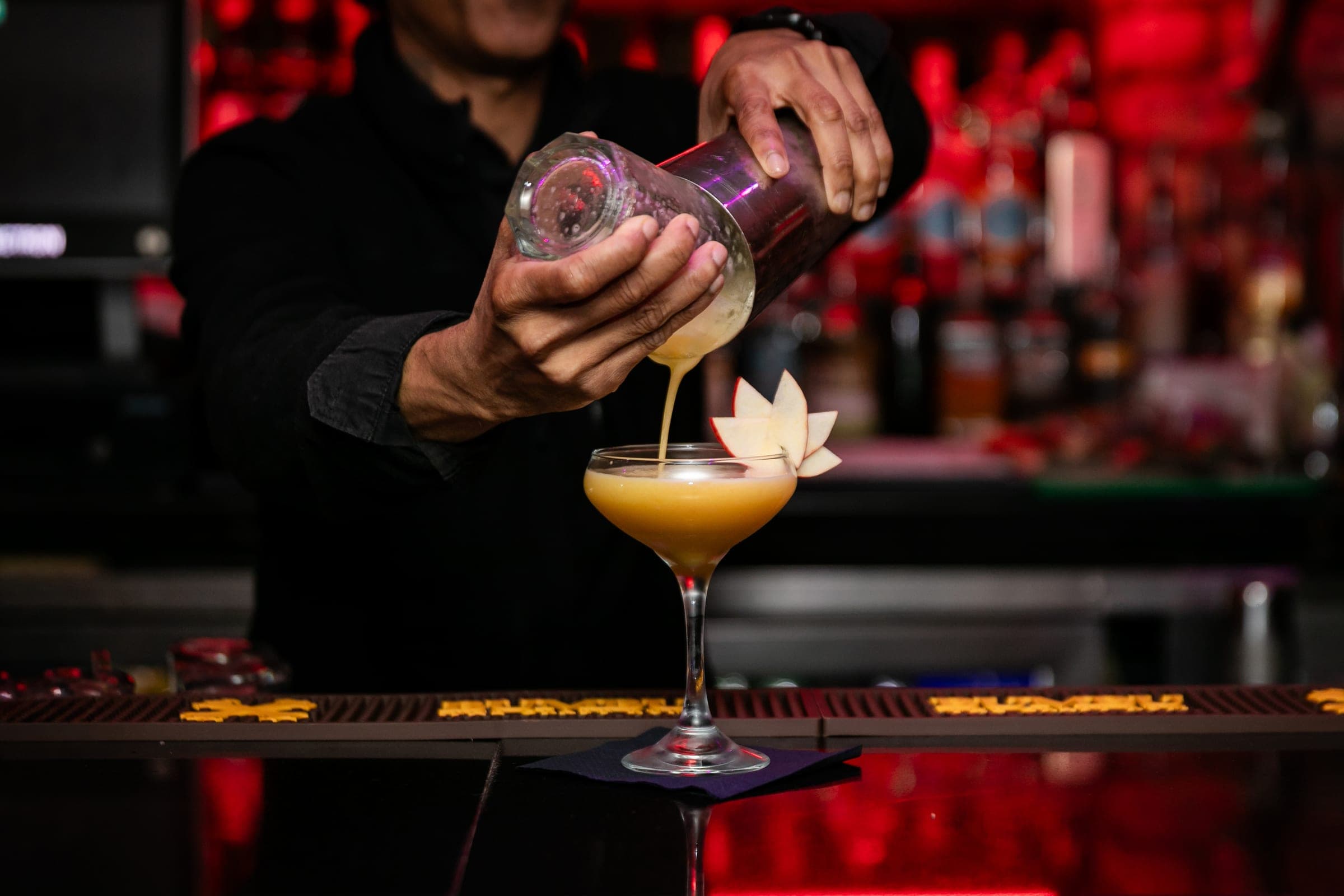 Bartender pouring a cocktail into a coupe glass at a bar lit in red