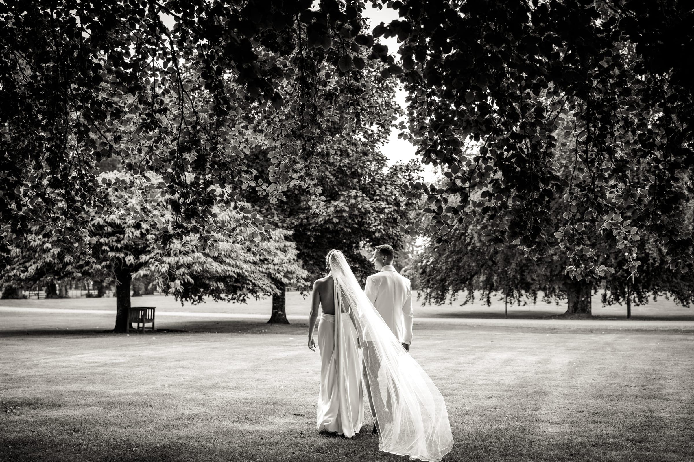Bride and groom walking away across a lawn under tall trees, her veil trailing behind, in black and white